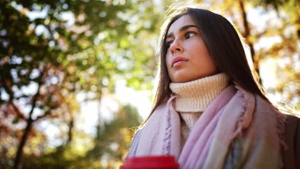 Portrait caucasian charming lovely dreamy lady hand hold glass coffee and enjoy drink in autumn park. Young woman look at nature landscape, yellowed leaves in trees against blue sky. Outdoors walk.