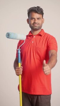 vertical shot of smiling painter with paint roller brush showing thumbs up sign by looking camera - concept of professional service, employment and home renovation.