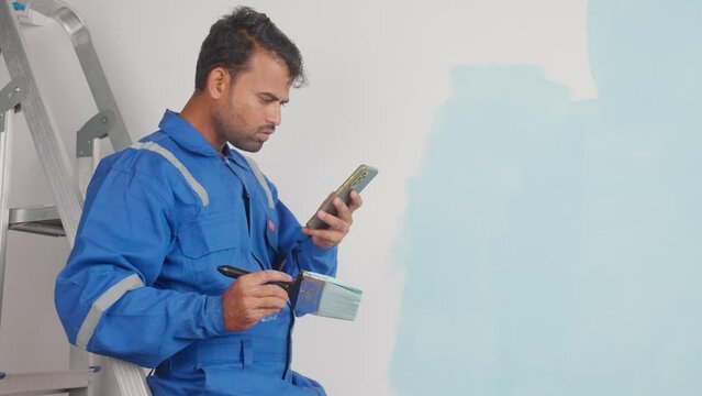 painter on ladder checking mobile phone while painting wall - concept of technology, skills and renovation