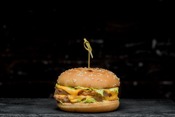 Burger with a latch on a wooden board on a black background;
bun sprinkled with sesame seeds; 
double cheese burger;