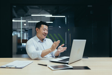 Fototapeta na wymiar Business meeting. A young asian male businessman conducts an online business meeting, conference. Sitting at the table in the office, looking at the laptop, applauding, thanking, smiling.