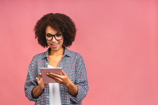 Portrait of young african american woman with curly african hair holding digital tablet and smiling standing over isolated pink background with copy space.