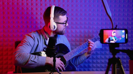 Young influencer playing guitar during podcast or live video streaming for audience from mobile phone at home - concept of art, hobby and video blog