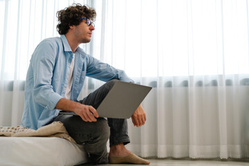 Man thinking some idea in bedroom. Businessman sitting on bed, browsing Internet, working with laptop at home