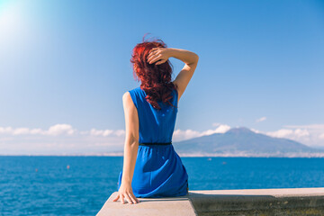 A young girl in a blue dress is sitting on the seashore, in the background the volcano Vesuvius Italy Naples