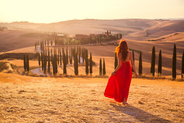 A girl at sunset in a red dress on a field in Italian Tuscany. Val d'Orcia. Beautiful landscape scenery at sunset of Tuscany in Italy
