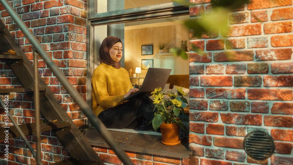 Poster creative muslim female in hijab working on a laptop computer while sitting on a window sill of her c