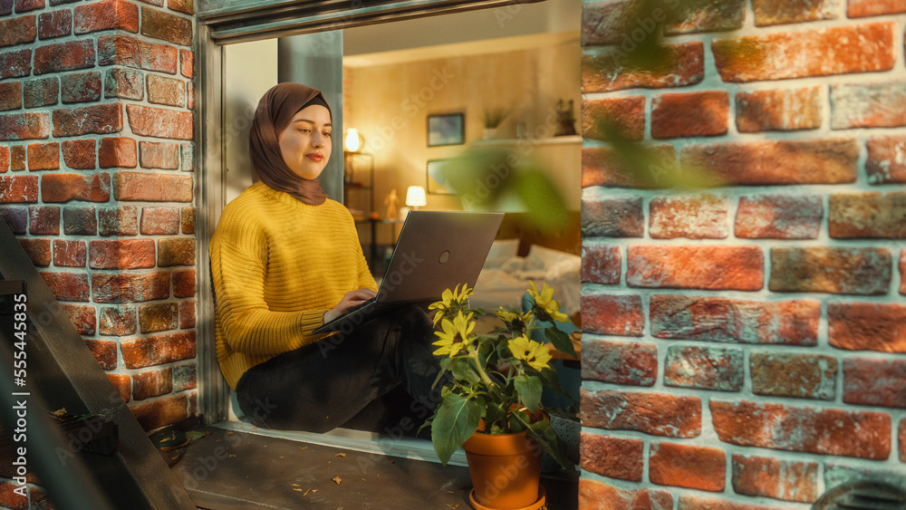 Aufkleber Young Muslim Female in Hijab Using Laptop Computer while Sitting on a Window Sill in Her Stylish Apartment. Creative Manager Working Remotely from Home. Arc Shot from Outside the Window.
