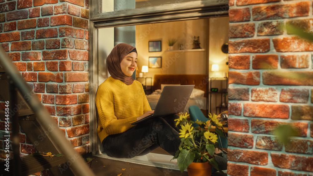 Poster creative muslim female in hijab working on a laptop computer while sitting on a window sill of her c