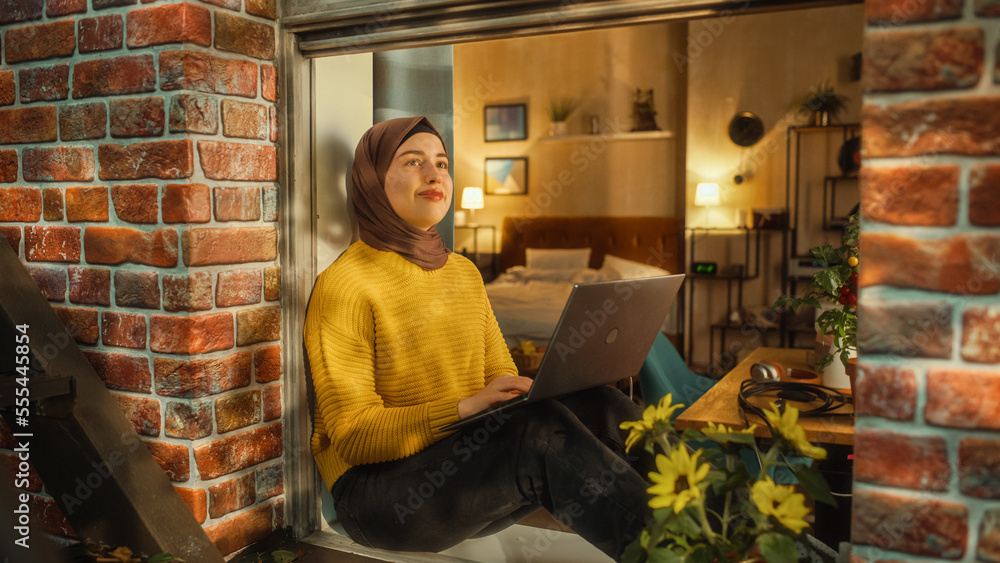 Aufkleber Young Muslim Female in Hijab Using Laptop Computer while Sitting on a Window Sill in Her Stylish Apartment. Creative Manager Working Remotely from Home. Arc Shot from Outside the Window.