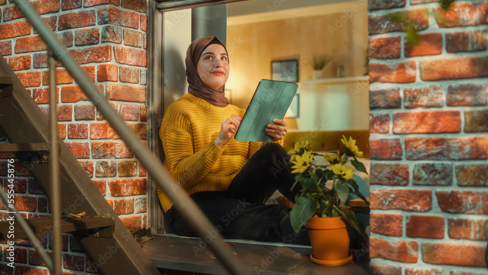 Poster muslim girl wearing a hijab, using a tablet computer while sitting on a window sill in brooklyn styl