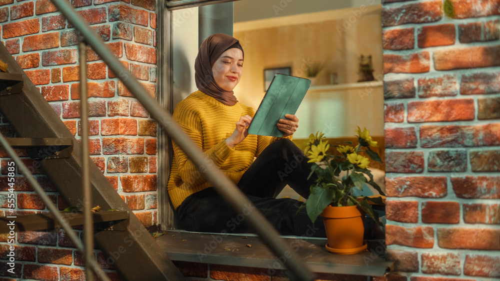 Aufkleber happy arab female in a hijab sitting on a window sill, checking social media, using online apps on t