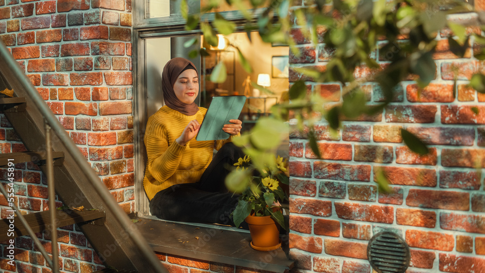 Poster portrait of a young arab woman using tablet computer while sitting on window sill in a multi-storey 