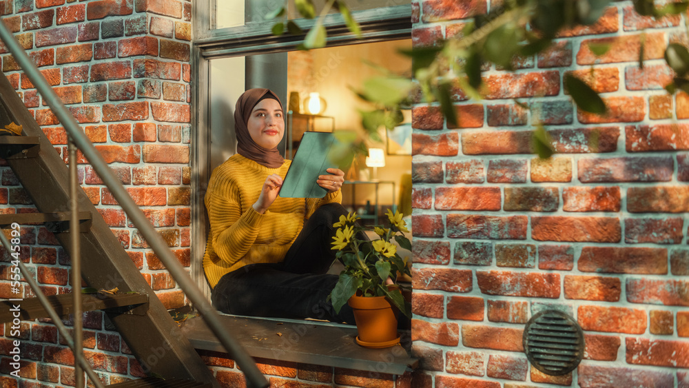Poster muslim girl wearing a hijab, using a tablet computer while sitting on a window sill in brooklyn styl