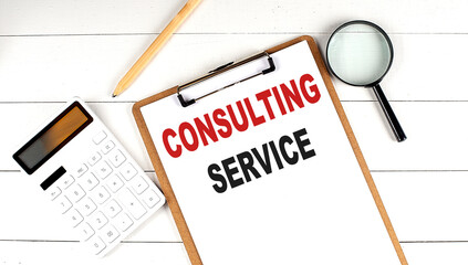 CONSULTING SERVICE words on clipboard, with calculator, magnifier and pencil on white wooden background
