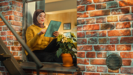 Portrait of a Young Arab Woman Using Tablet Computer while Sitting on Window Sill in a Multi-Storey...
