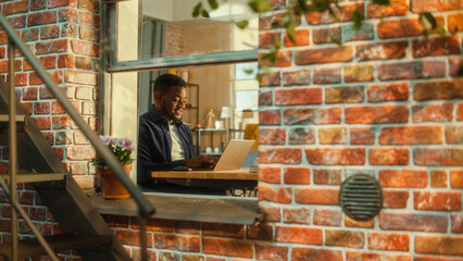 Young Handsome Male Using Laptop Computer, Sitting in Living Room in Apartment. African American Businessman Checking Social Media, Smiling, Answering Emails from Home. Inside Apartment Window View.