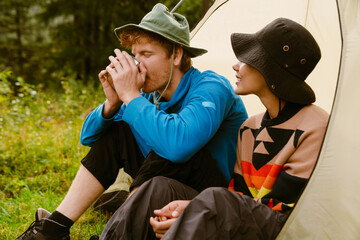 Young man drinking tea while sitting with his girlfriend at camping tent