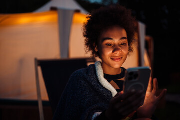 Smiling african woman using mobile phone while sitting on chair near glamping tent