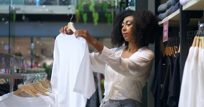 Pleasant milti-ethnic young pretty woman examining a white t-shirt while shopping for new garment, in fashion store. Sale shopping fashion and people concept