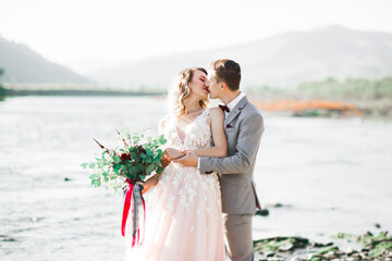 Elegant stylish happy blonde bride and gorgeous groom on the background of a beautiful river in the mountains