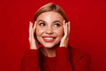 Smiling blonde woman touching her lips while standing isolated over red wall