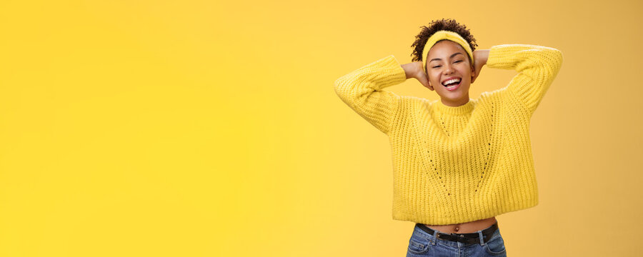 Carefree joyful lucky african-american female student in sweater headband lay back hands behind head relaxed chilling pose having day-off weekend smiling broadly laughing, yellow background
