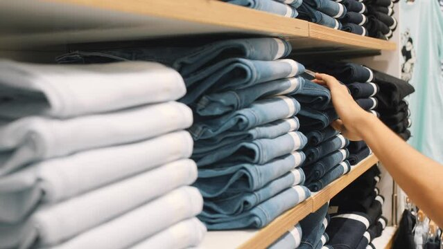 Young Woman Taking Pair of Blue Denim Jeans From Stacks in Clothing Shop. Female Hands Choosing Correct Pants Size in Shopping Mall. 4K Consumerism Concept.