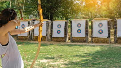 Fototapeta premium Hands of woman aims archery bow and arrow to colorful target in shooting range during training. Exercise and concentration with outdoor archery. Selective focus on hand. Sport and Recreation concept.