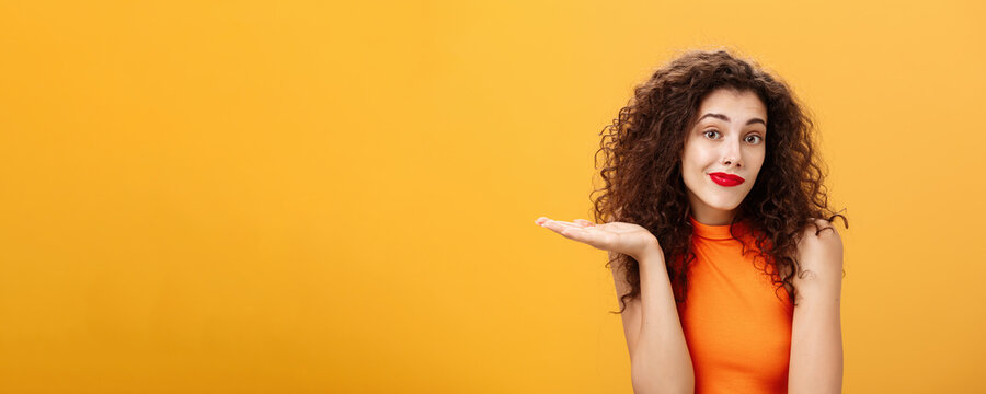 Well sorry. Unaware and clueless silly charming woman with curly hairstyle shrugging and smirking with uncertain look raising palm in who knows gesture, posing uninvolved over orange background