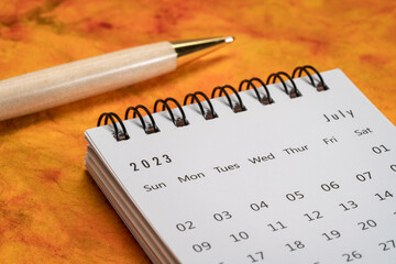 July 2023 - small spiral desktop calendar with a stylish pen  against marbled paper, time and business concept