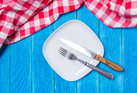 Top view background with squared empty white ceramic plate on vintage weathered blue wooden boards and red checkered kitchen towel on top.