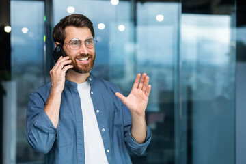 Successful smiling businessman in casual shirt talking on phone, male boss in glasses and beard near window inside modern office.