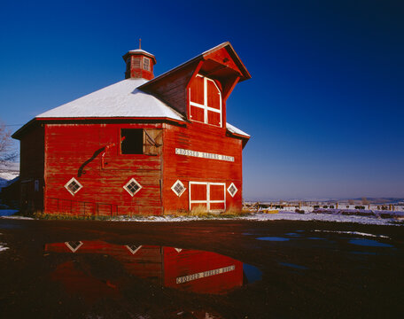 Agriculture - Crossed Sabres Ranch red octagonal barn with snow on the ground around it and a reflection in a puddle / Joseph, Oregon, USA.