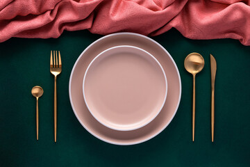 Festive place setting with pink napkin. Empty plates and gold cutlery on dark green background. Top...
