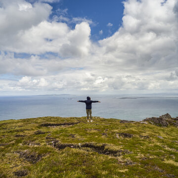 A woman stands with outstretched arms and looks out at the ocean and coastline while hiking at Brandon Point, Dingle Peninsula; Castlegregory, County Kerry, Ireland