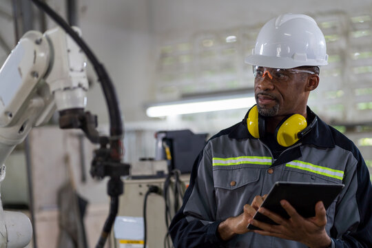 African American Male Engineer Worker Holding Digital Tablet Working With Welding Robot Arm In Workshop. Male Worker Control Automatic Welding Robot Arm In Factory