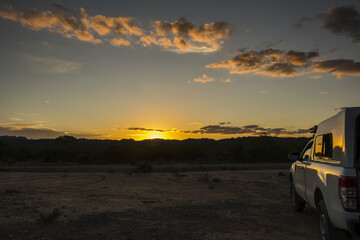 The sun is setting behind a range of hills, lighting up the few clouds in orange and red hues. The side of a camper truck is visible on the right hand side of the image; San Luis, Argentina