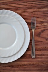 A white plate on the left and a fork on the right, dinnerware on a brown textured wooden table