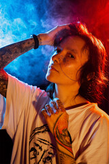 Stylish young tattooed woman with face piercing posing on  dark background with smoke in neon...
