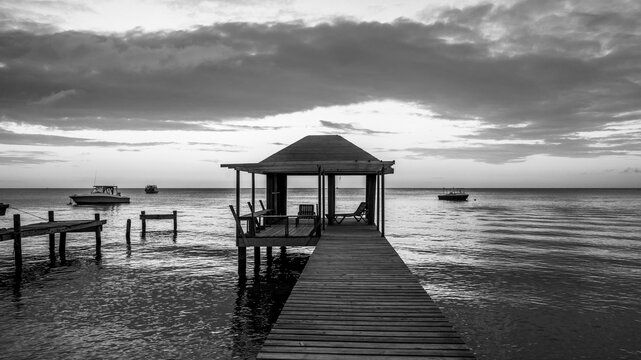 Black and white image of a dock with seating and boats mooring on the tranquil water at sunset; Roatan, Bay Islands Department, Honduras