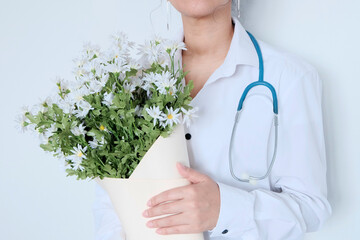 The doctor is young, an Asian girl with black hair in medical white clothes with a stethoscope around her neck. Holds a bouquet of  chamomile flowers in her hands. Light background. Medic day 