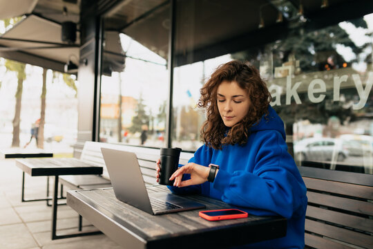 Stylish pretty European girl with fluffy hair wearing blues shirt sitting outdoor in city cafe, looking at smartwatch, drinking coffee and working on laptop