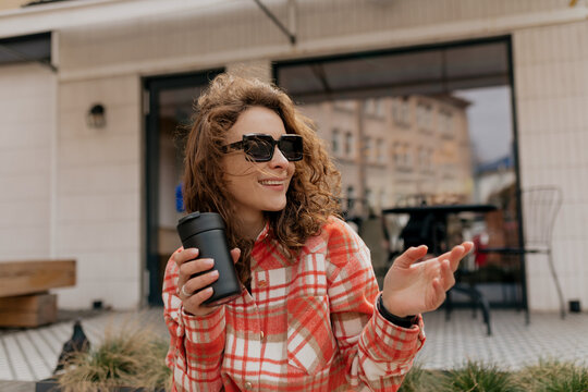 Stylish european incredible girl with curly hair in sunglasses and bright checkered shirt drinking coffee and looking aside outdoor 