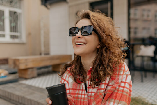 Lovely cute happy girl with wavy hair and wonderful smile in sunglasses enjoying sunny weather in the city while drinking coffee 