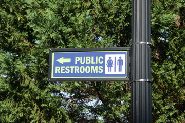 Public Restrooms Sign on a black pole with evergreen tree in background.