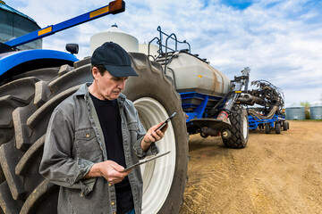 Farmer using a smart phone and tablet while standing on a farm beside equipment; Alberta, Canada