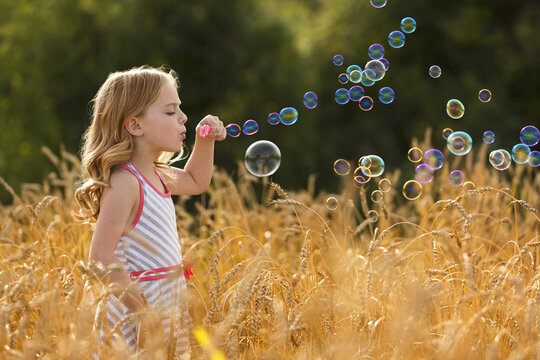 Young girl stands in a golden wheat field blowing bubbles; Alberta, Canada