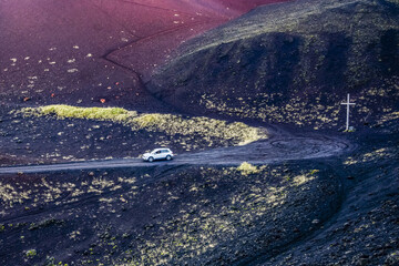 A car travels on a road on black volcanic landscape with a cross on the roadside on one of the Westman Islands; Vestmannaeyjar, Southern Region, Iceland
