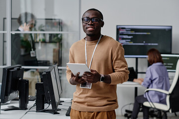 Portrait of African American IT specialist in eyeglasses smiling at camera while working online on...